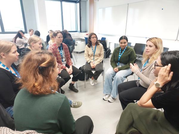 Women in a mentoring circle talk in a classroom; participants with blue lanyards sit in chairs facing each other.