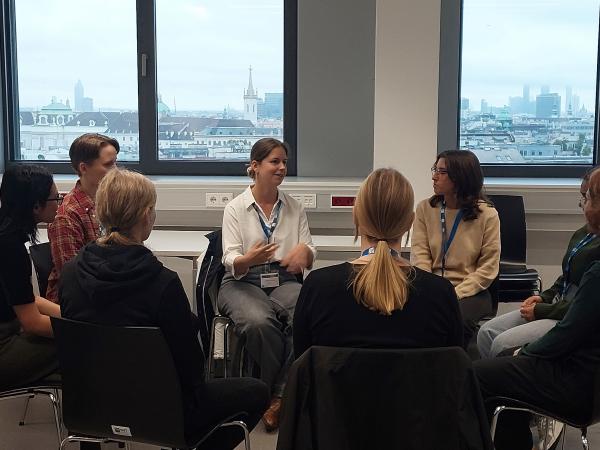 Mentoring circle in a classroom; a facilitator speaks to women seated in a ring, city skyline visible through large windows.