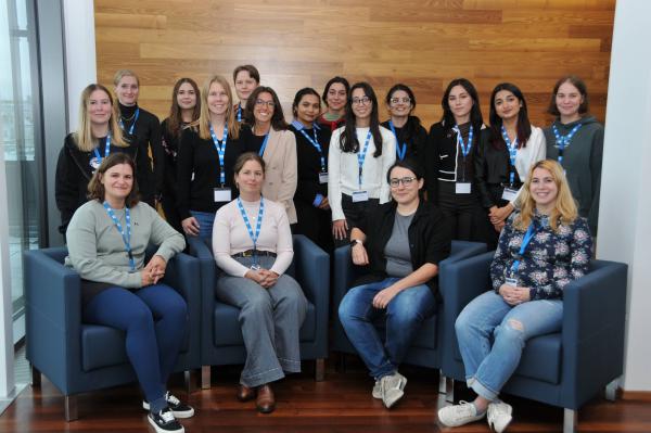 Indoor group photo of female participants at the Summer School on Artificial Intelligence and Cybersecurity; four speakers are seated in blue armchairs, with others standing against a wooden wall.