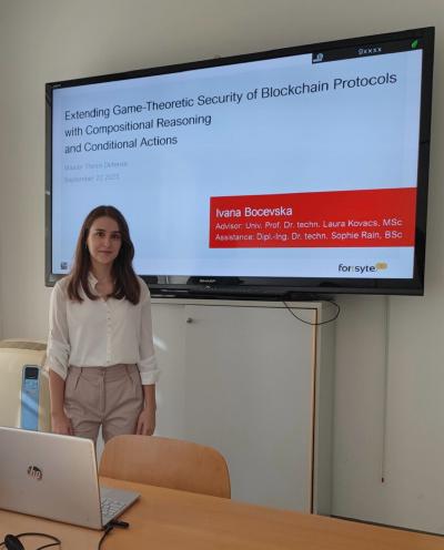 A presenter stands beside a table with a laptop and cables, speaking to an audience. Behind her, a wall-mounted screen shows a slide of titled “Extending Game-Theoretic Security of Blockchain Protocols with Compositional Reasoning and Conditional Actions.”