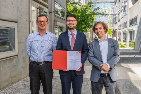 Three men stand outside a modern academic building. The man in the center holds a red folder displaying an official certificate and smiles at the camera. The image captures a celebratory moment following a PhD defense.