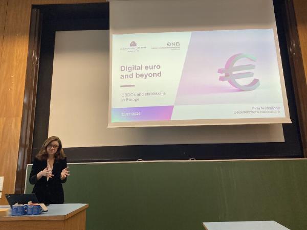 A woman stands at the front of a lecture hall, speaking and gesturing with her hands beside a projected slide. A green chalkboard, classroom desks, and a table with a laptop are visible in the foreground.