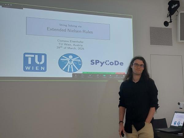 Clemens Eisenhofer is standing in front of a presentation screen that displays the title of the presentation and the logos of TU Wien and SPyCoDe.