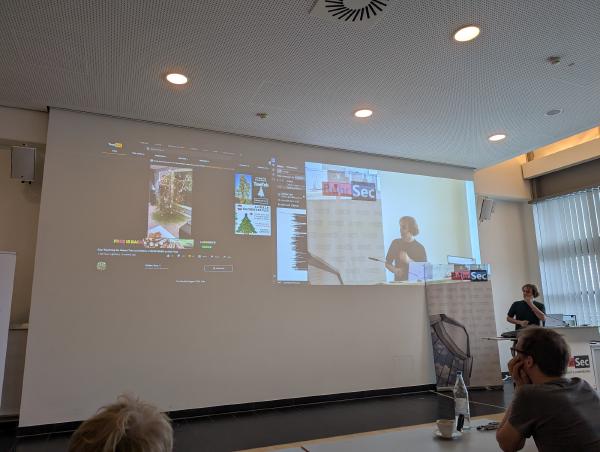 The image captures a professional conference setting at the RuhrSec conference with Daniel Gruss as the speaker. The main projection screen displays a variety of browser tabs and content related to statistics. He is also shown on a smaller screen to the right, indicating a live presentation. Attendees are visible in the foreground, listening attentively, with one person’s head prominently in focus.