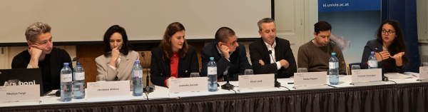 A panel discussion with seven participants seated behind a long table. The background includes a projection screen and a blue banner featuring the University of Vienna logo. One panelist is actively speaking and using hand gestures, while the others appear to be listening attentively.