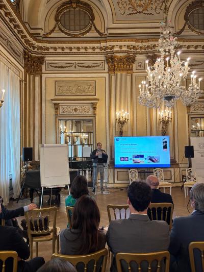 A speaker is presenting to an audience in an elegant, ornately decorated room with high ceilings, large mirrors, and intricate molding. A large screen beside the speaker displays a slide with text and images.