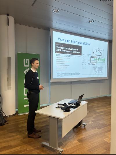A young man dressed in business casual attire stands next to a white desk in a modern conference room, giving a presentation. Behind him, a large projection screen displays a slide titled 'Was sind Internetausfälle?' ('What are internet outages?') with a diagram and a list of major internet outages in 2024.