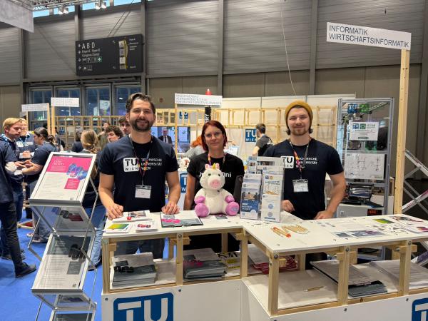 Martina Lindorfer stands with two other TUW representatives behind a counter displaying various informational materials. The booth features posters and pamphlets related to cybersecurity and engineering programs offered by TU Wien. The environment suggests a lively event with other attendees and booths in the background.