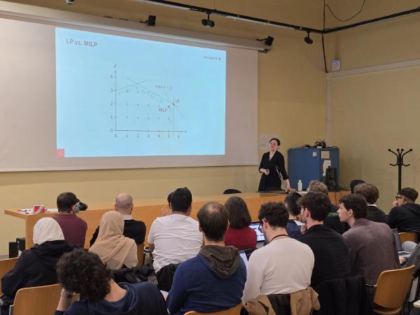 Maria Eichlseder is giving a presentation in a lecture hall to an audience of students. She stands near a desk with a microphone and laptop, while the audience listens attentively, with some taking notes on laptops. A slide titled “LP vs. MILP“ is projected on the screen, showing a graph comparing linear programming (LP) and mixed-integer linear programming (MILP). The setting appears academic and professional.