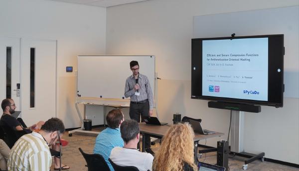 Stefano Trevisani presenting at a professional conference, standing next to a digital screen displaying a presentation. The audience, comprised of various attendees focused on the speaker, is seated at tables in a modern, well-lit room.