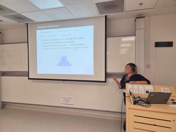 Elena Andreeva stands in a classroom giving a presentation. On the screen is a slide titled “ButterKnife mIFI Instance,” containing mathematical formulas, bullet points, and a diagram. She is gesturing toward the projection while explaining the content.