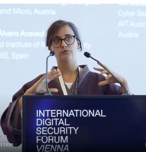 Johanna Ullrich is speaking at a podium. The podium displays the text ‘INTERNATIONAL DIGITAL SECURITY FORUM VIENNA.‘ The background shows blurred text projected on a screen