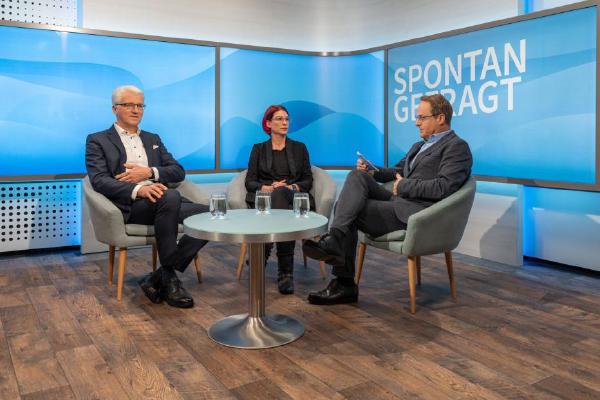The image shows a television talk show setting with three participants - two men and a woman. They are positioned around a round table in a studio with a blue-themed background that reads 'SPONTAN GEFRAGT'.