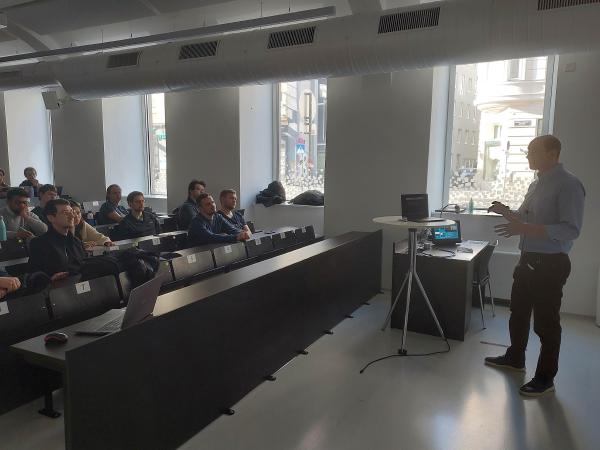 A lecturer is giving a presentation in a modern classroom with large windows. He is gesturing with his hands while speaking to an audience of students seated in tiered rows. The audience, consisting mostly of young adults, is attentively listening, some with laptops open.