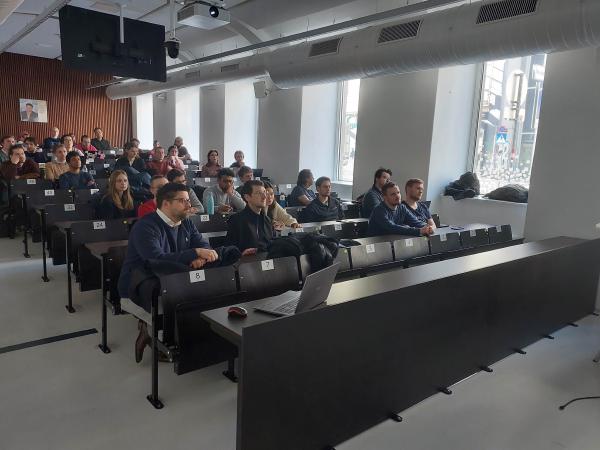 A diverse audience of students and professionals is seated in a modern lecture hall, attentively listening to a presentation. Some attendees have laptops open, while others are taking notes or observing the speaker.