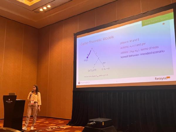 A presenter speaks beside a large slide titled “Game-Theoretic Models” in a conference room