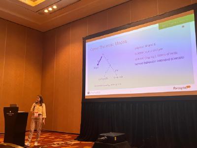 A presenter speaks beside a large slide titled “Game-Theoretic Models” in a conference room A presenter speaks beside a large slide titled “Game-Theoretic Models” in a conference room
