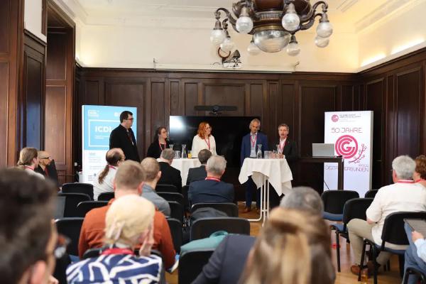 Panel discussion taking place in a formal conference room. Five panelists stand behind two high tables, engaged in conversation. Audience members sit facing the panel, listening attentively. Two vertical banners are visible, one featuring the ICDL logo and the other celebrating the 50th anniversary of the Österreichische Computer Gesellschaft.