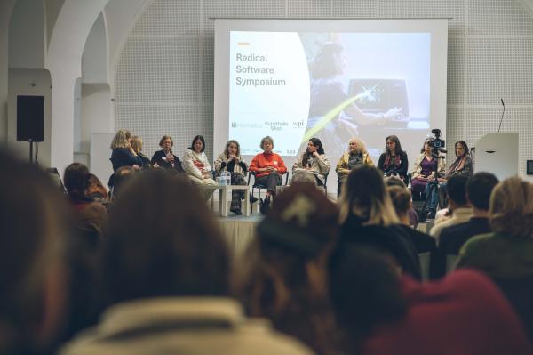 A panel of eight diverse women sits on a stage at the 'Radical Software Symposium' in a modern venue. A large screen behind them displays a presentation related to the event, and the session appears well-attended by an engaged audience.