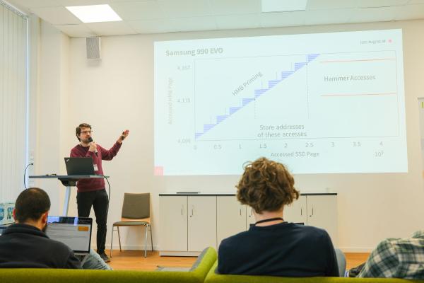 Jonas Juffinger stands at the front, beside a laptop, gesturing towards a graph displayed on the screen behind him, which details data related to “HMB-based SSD Rowhammer“.
