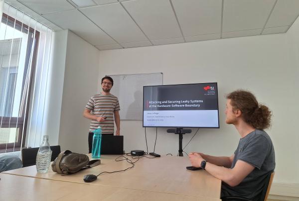 Jonas Juffinger is presenting in a bright conference room. He stands next to a large screen displaying a presentation slide titled 'Attacking and Securing Leaky Systems at the Hardware-Software Boundary.' Another man sits at the table, listening attentively.