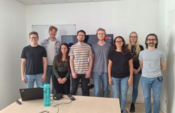 A group photo of eight people standing in a row inside a conference room.